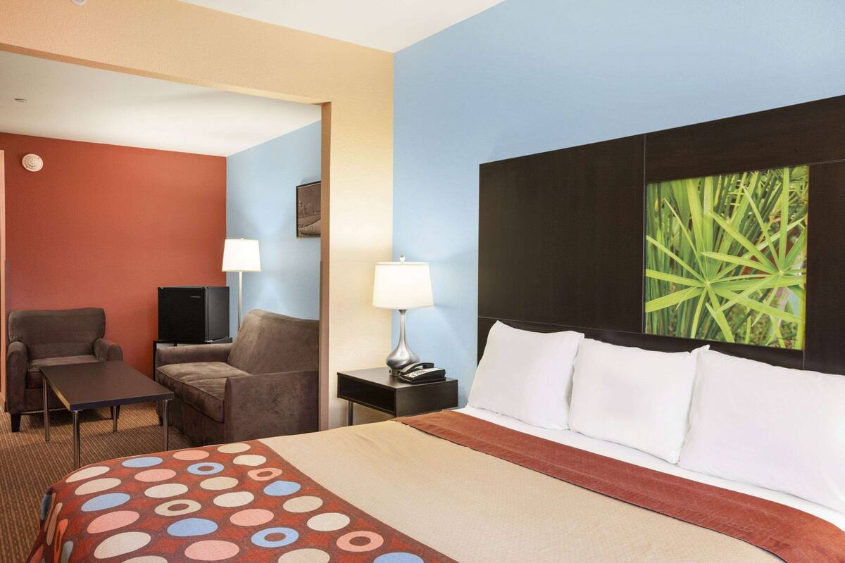 A hotel room with a bed, two armchairs, a small table, a TV, a nightstand with a lamp, and a wall art piece.