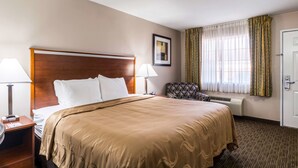 Habitación de hotel con una cama grande, dos mesitas de noche con lámparas, una silla, un cuadro enmarcado en la pared y una ventana con cortinas.