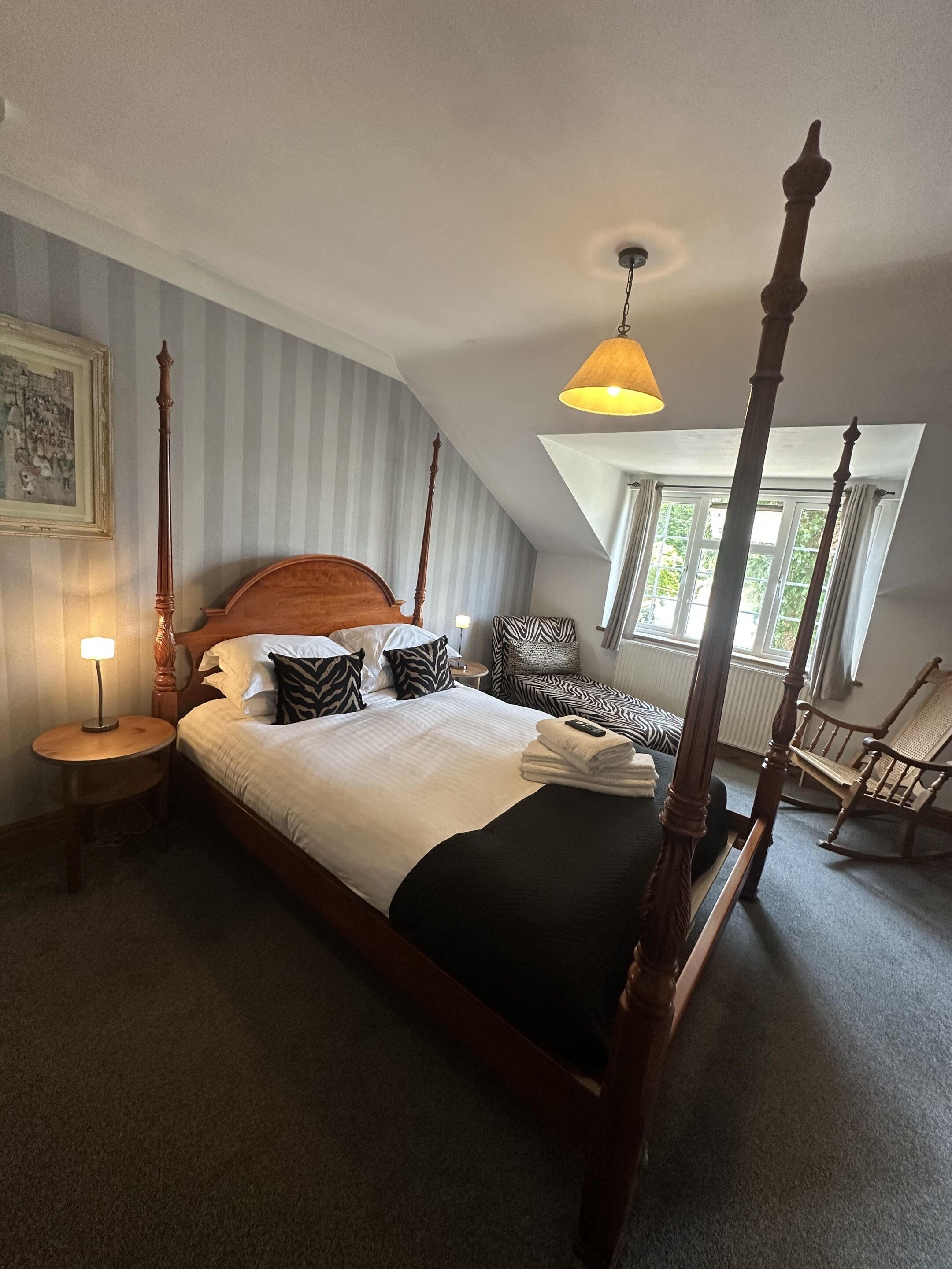 A four-poster bed with a wooden headboard, a round bedside table with a lamp, a zebra-patterned pillow, and a chair with a woven seat.