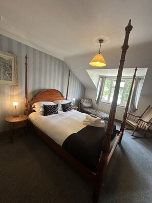 A four-poster bed with a wooden headboard, a round bedside table with a lamp, a zebra-patterned pillow, and a chair with a woven seat.