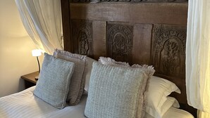 Un lit avec du linge de lit blanc et deux oreillers gris, une tête de lit en bois ornée de motifs sculptés et un baldaquin blanc.