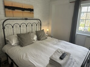 A neatly made bed with white linens and grey pillows, a remote control on a folded towel, and a painting of a sunset on the wall.
