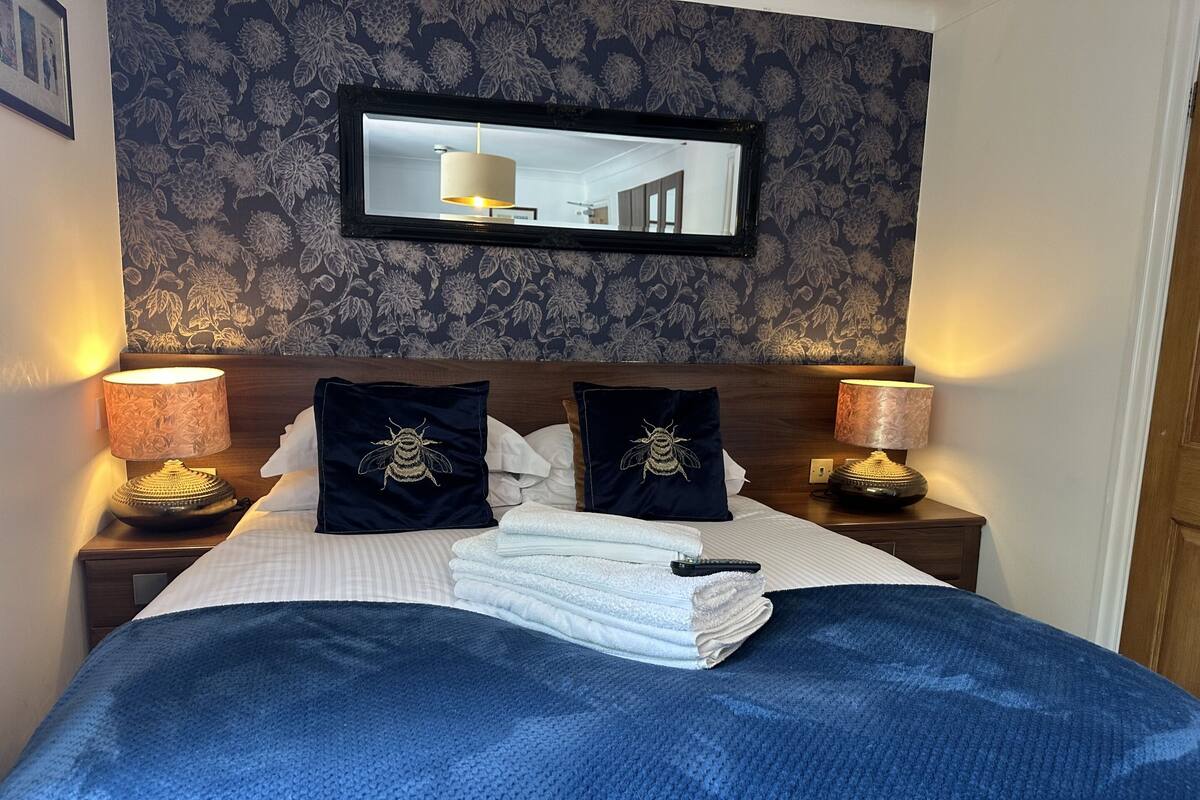 A neatly made bed with a blue blanket, two pillows with bee embroidery, and a stack of white towels. There are two bedside lamps with floral designs and a mirror above the bed.