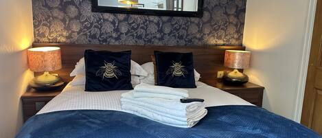 Ein ordentlich bezogenes Bett mit einer blauen Decke, zwei Kissen mit Bienenstickerei und einem Stapel weißer Handtücher. Zwei Nachttische mit floralem Dekor und ein Spiegel über dem Bett sind ebenfalls vorhanden.
