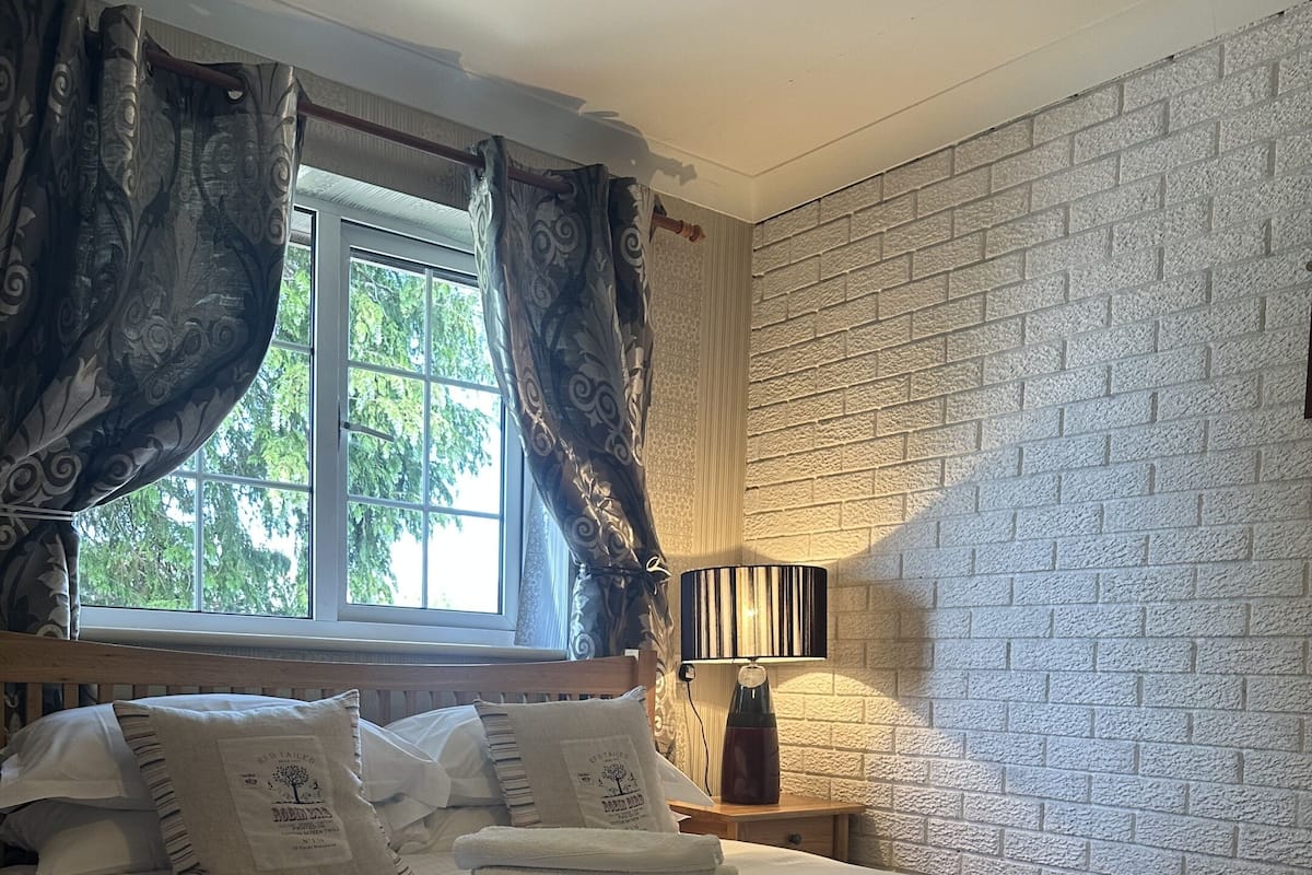 A neatly made bed with white linens and a set of folded towels, a bedside lamp, and a window with curtains.