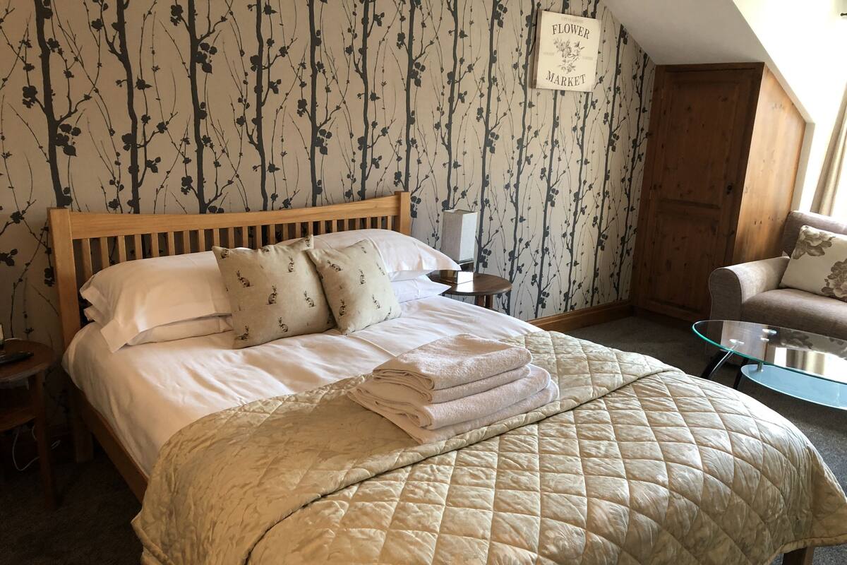 A neatly made bed with a quilted cover, a wooden headboard, and matching pillows. There is a small table with a glass top beside the bed.