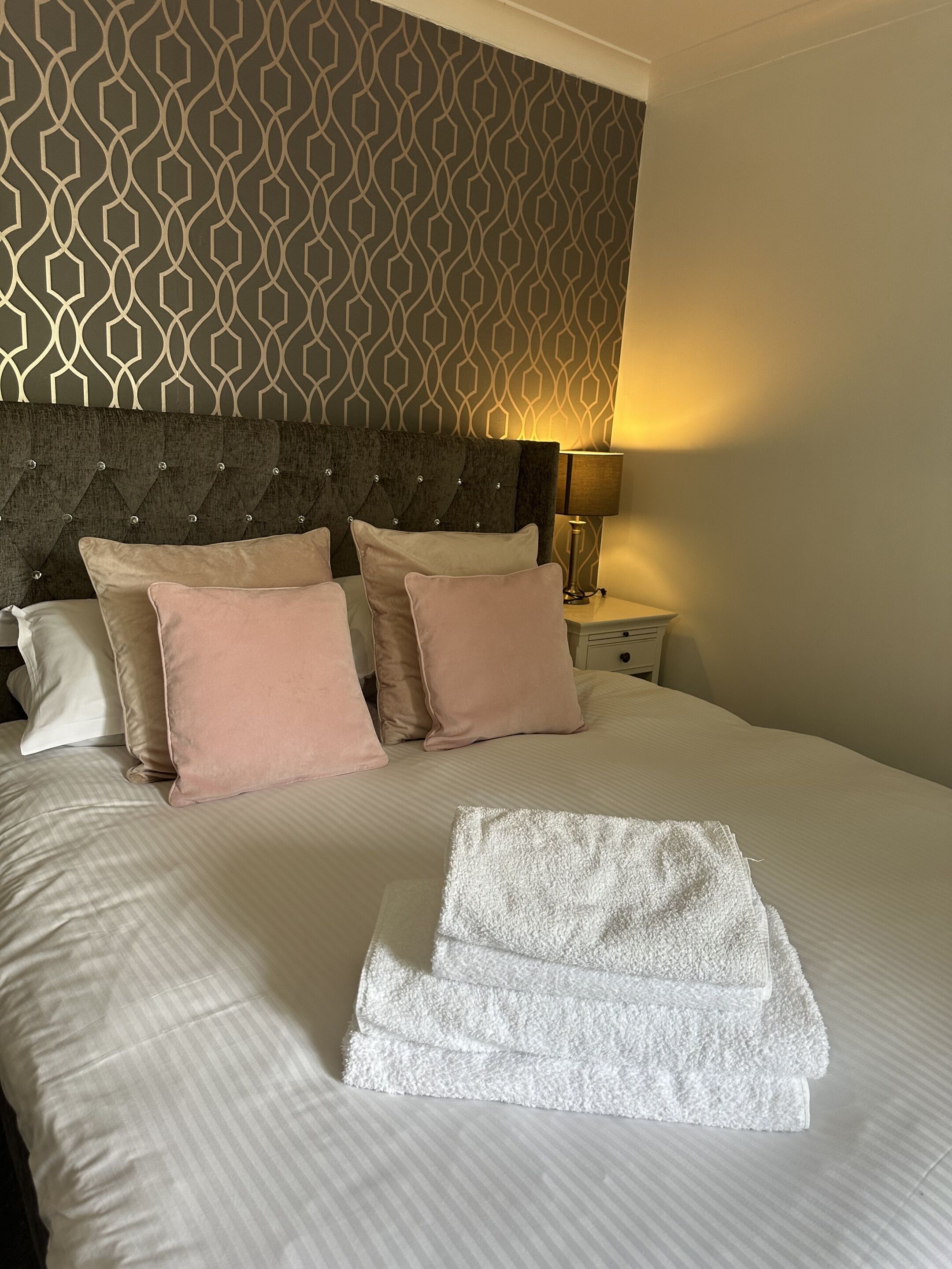 A neatly made bed with pillows and a folded towel, a headboard, a bedside lamp, and a patterned wall.