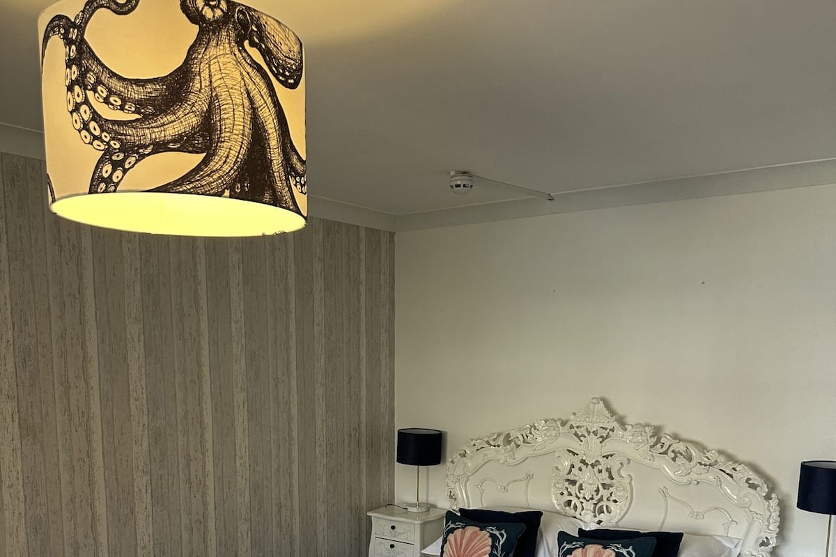 A neatly made bed with a white headboard and decorative pillows, a bedside table with a lamp, and a wall with vertical stripes.