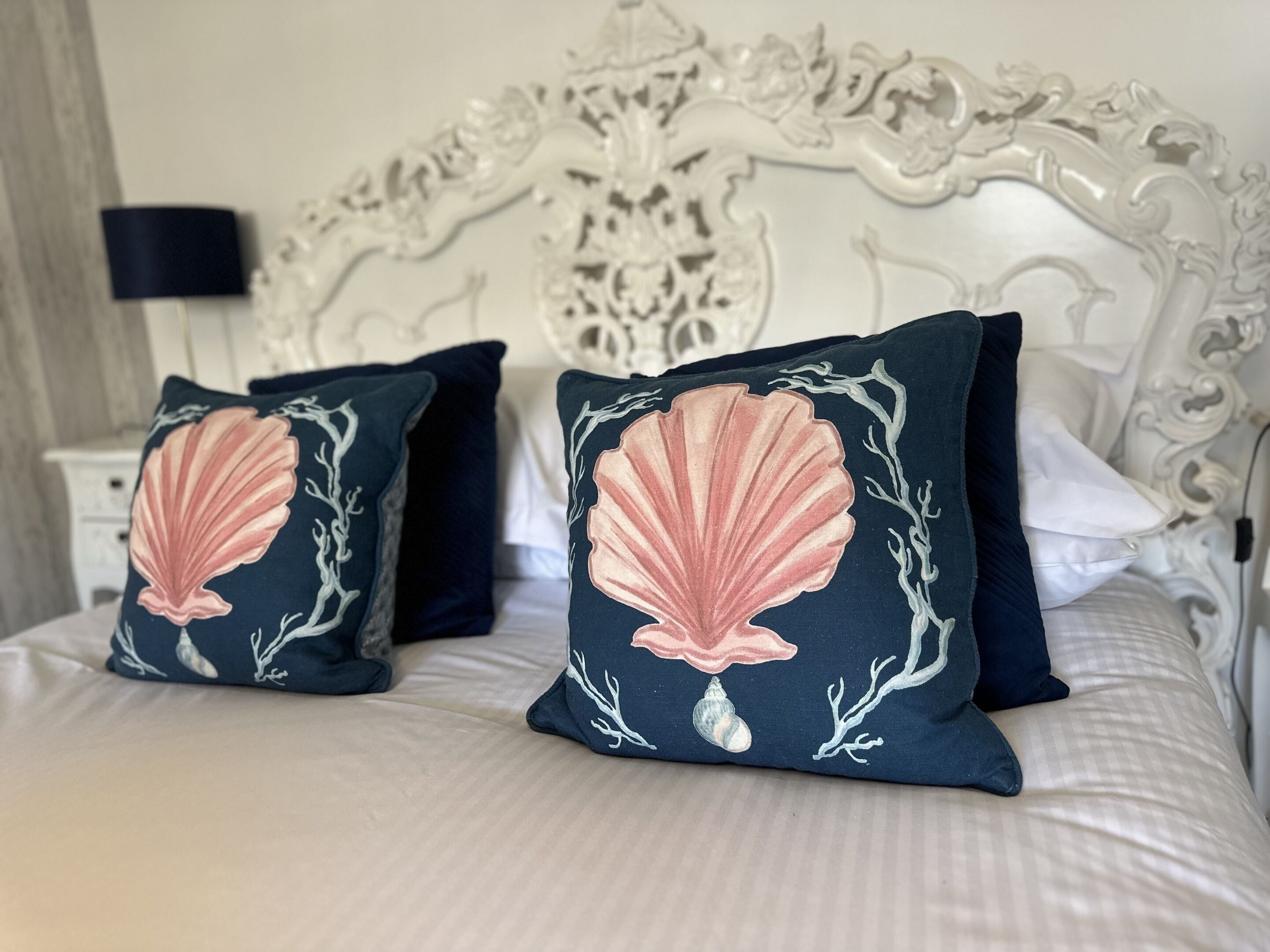 A bed with two navy pillows featuring a pink shell design, a white ornate headboard, and a bedside table with a lamp.