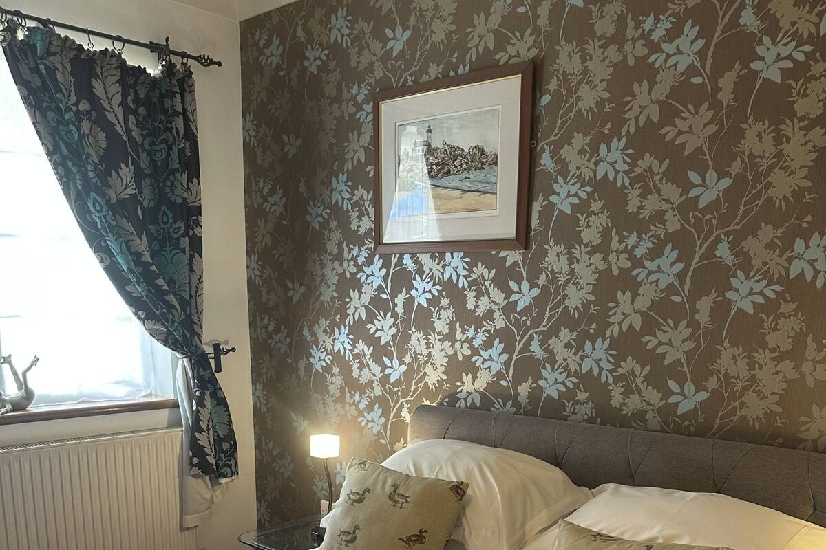 A neatly made bed with two pillows, a window with curtains, a radiator, and a framed picture on the wall.