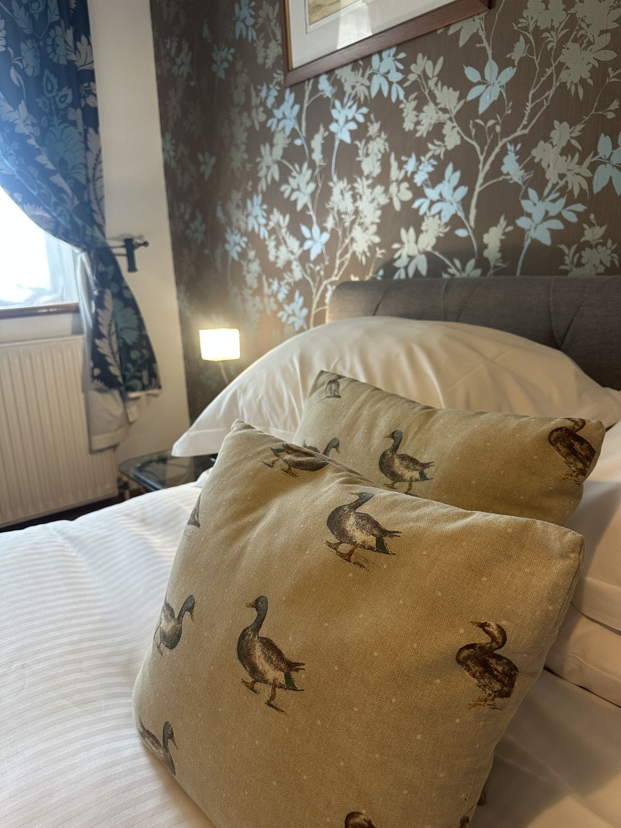 Een bed met twee kussens met een eendendesign, bloemenbehang en een nachtkastje met een lamp.