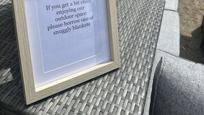 Une pancarte encadrée portant un message concernant l’emprunt de couvertures, placée sur une table en osier dans un cadre extérieur, avec une architecture en pierre en arrière-plan.