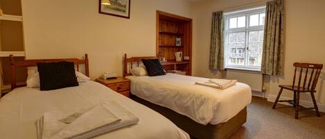 A hotel room with two beds, a wooden nightstand, a bookshelf, a window with curtains, and a chair.