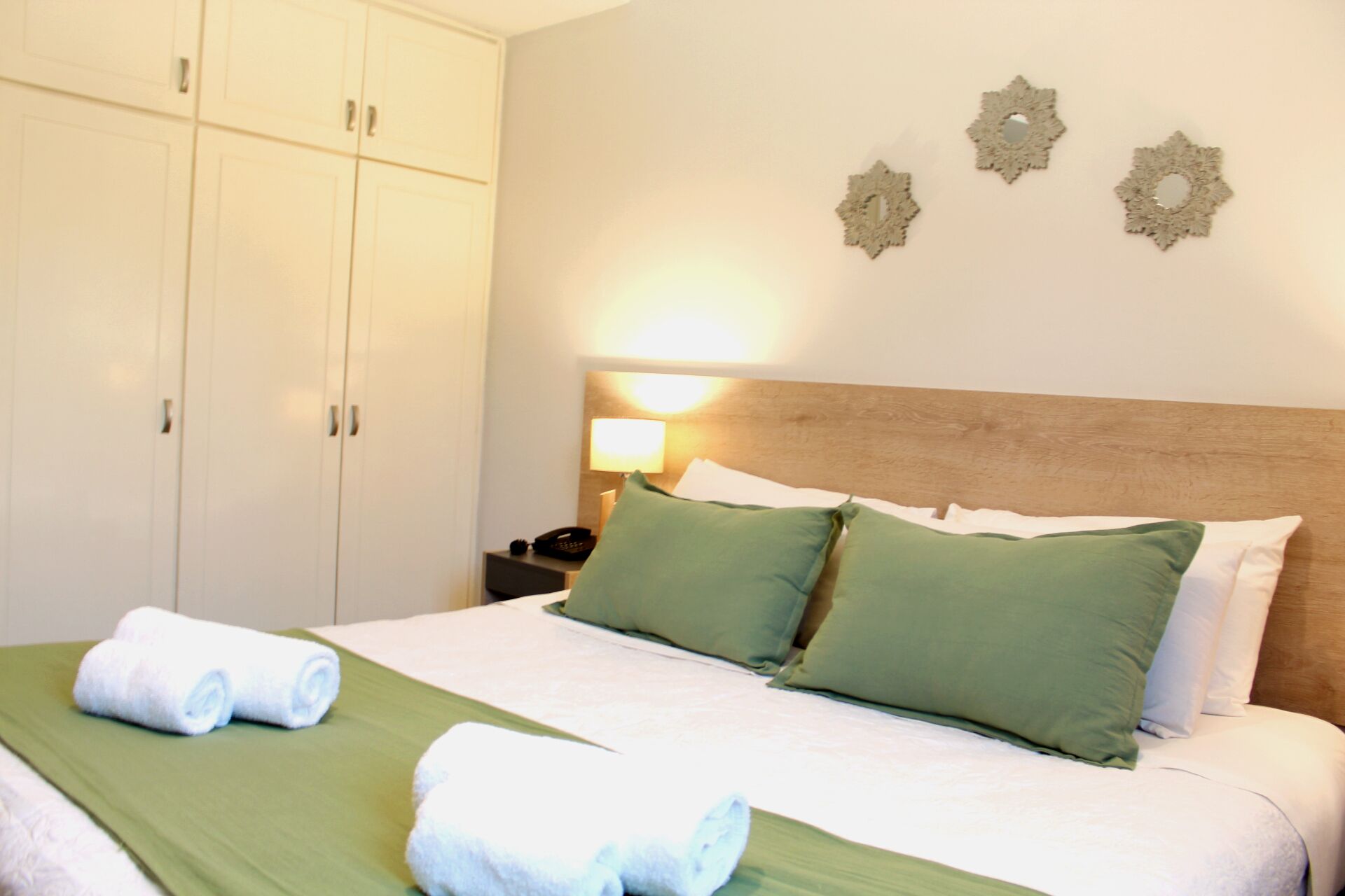 A hotel room with a bed, green bedspread, white pillows, and a nightstand with a lamp.