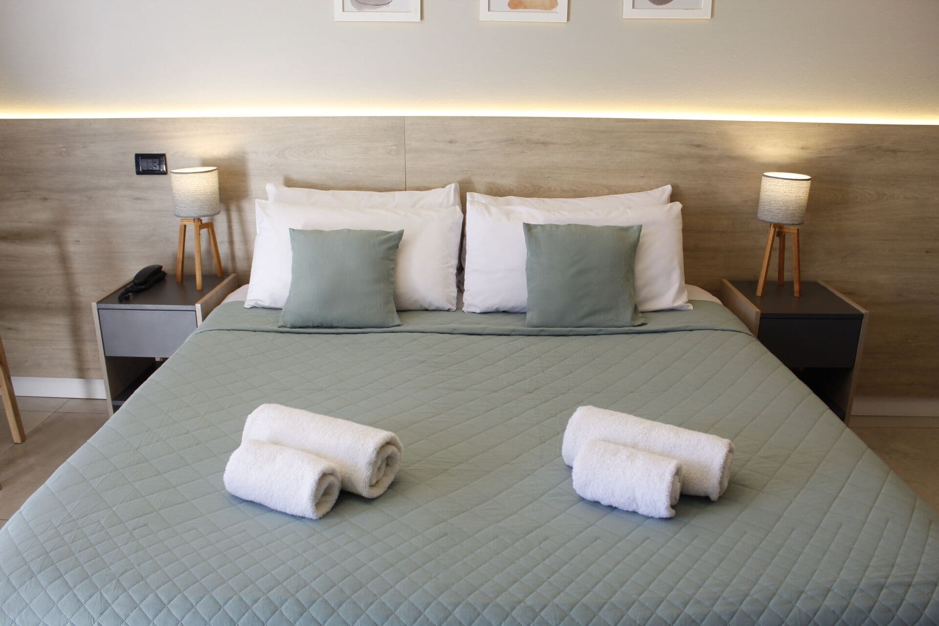 Un letto rifatto con una coperta verde trapuntata, due cuscini bianchi e due asciugamani bianchi arrotolati. Sono presenti due comodini con lampade, uno scollettato e un termostato a parete.