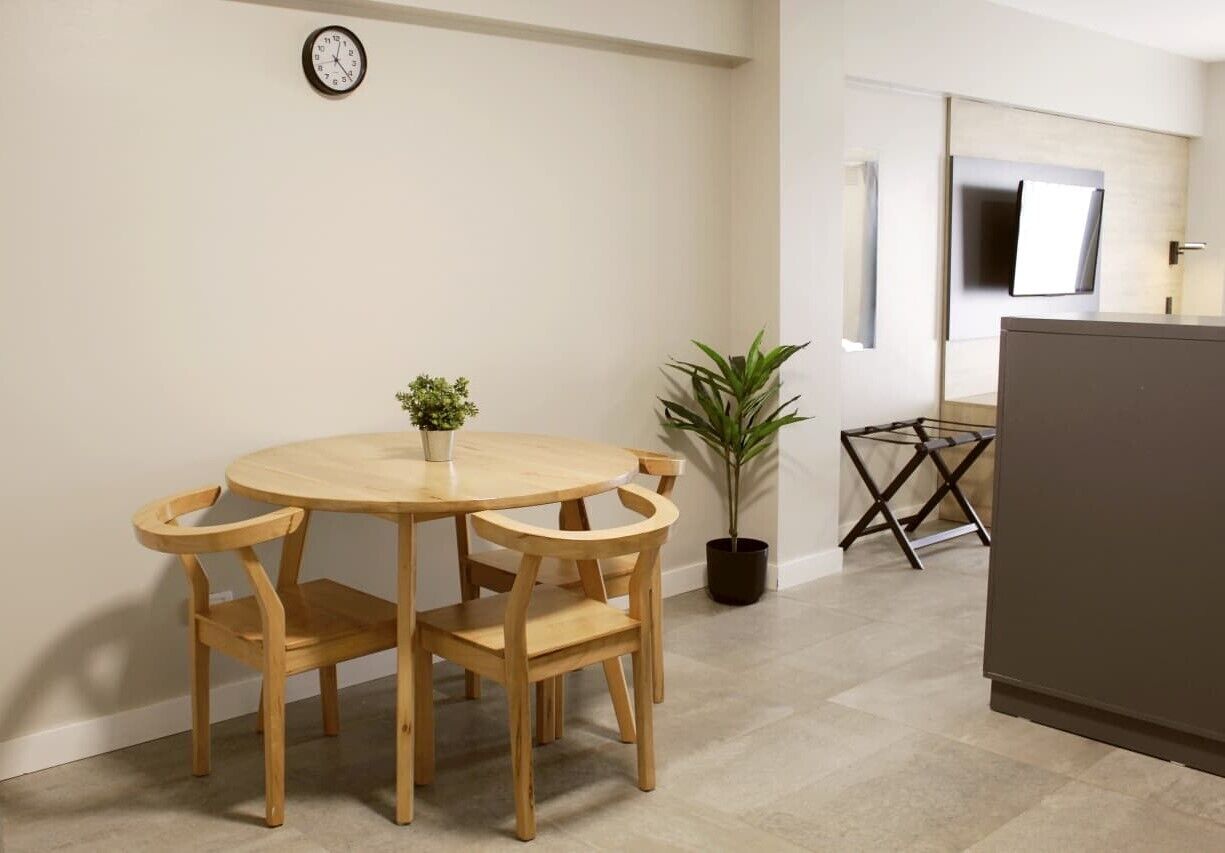 A room with a round wooden table and chairs, a potted plant, a small clock on the wall, and a television mounted on the wall.