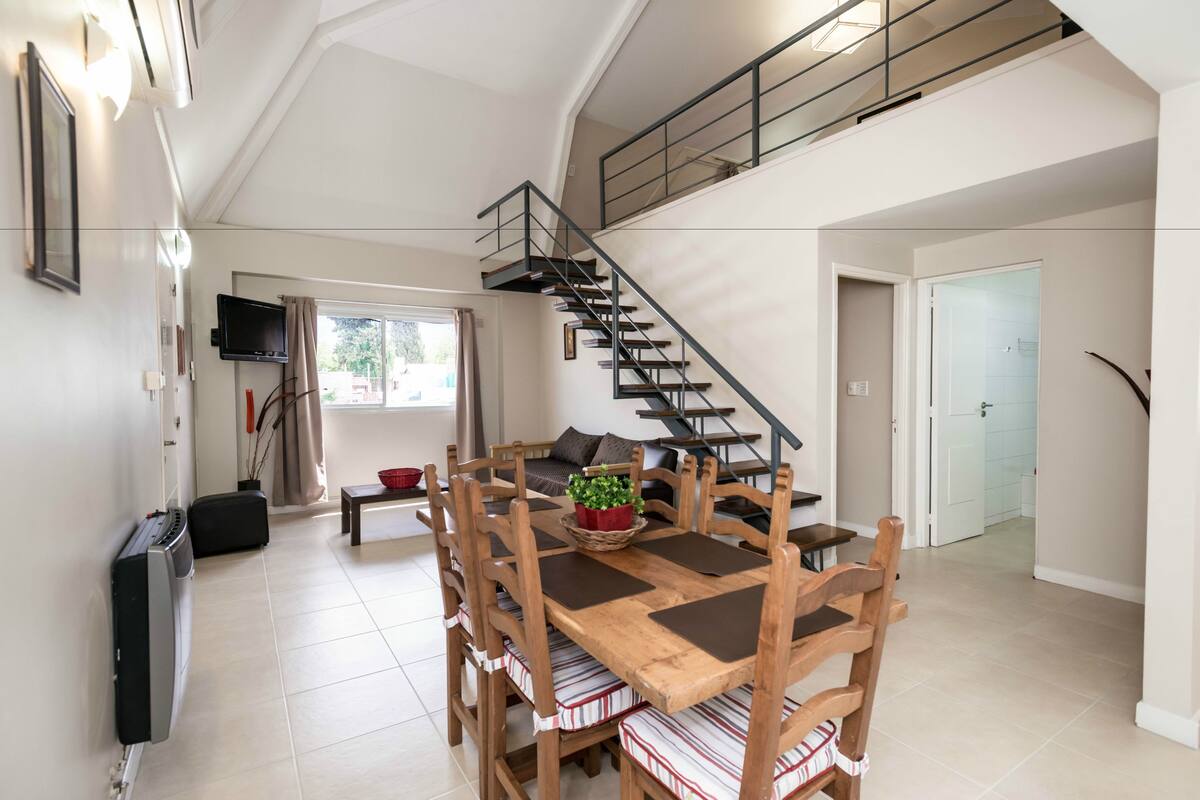 Amplia sala de estar con una mesa de comedor de madera, sillas, una escalera y un televisor montado en la pared.