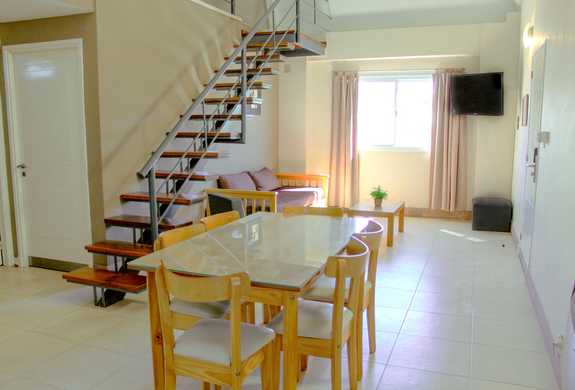 Un coin repas avec une table à plateau de verre et des chaises en bois, un escalier avec des rampes en métal, un canapé, une petite table avec une plante et un téléviseur fixé au mur.