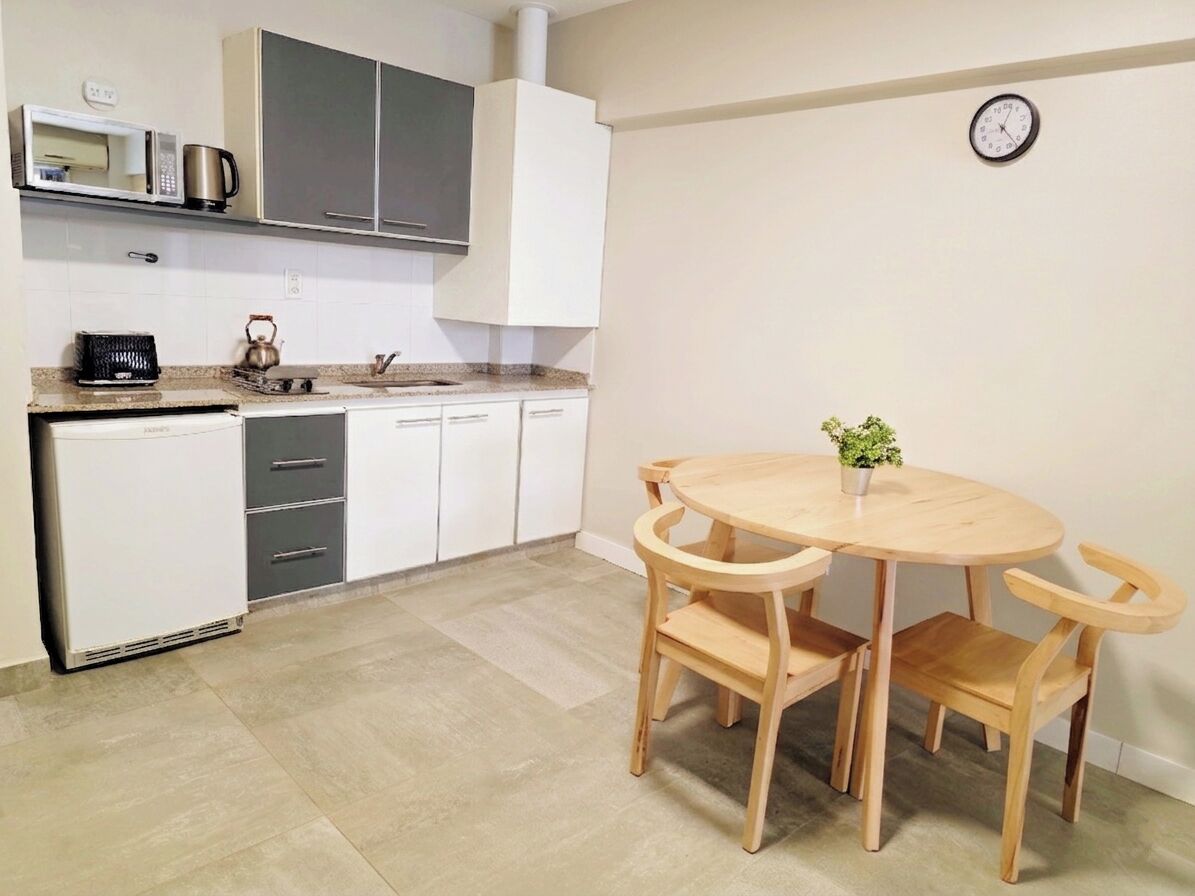 Una cocina compacta con armarios blancos, microondas, hervidor y una pequeña mesa de comedor con sillas.