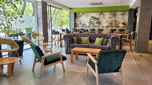 Un espace de détente intérieur moderne, avec un mélange de chaises et de canapés, dont un grand canapé moelleux agrémenté de coussins verts, et une fresque représentant des plantes tropicales sur le mur.