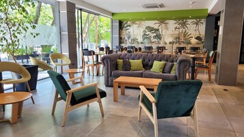 Un área de estar interior moderna con una variedad de sillas y sofás, incluyendo un sofá grande y acolchado con cojines verdes, y un mural de plantas tropicales en la pared.