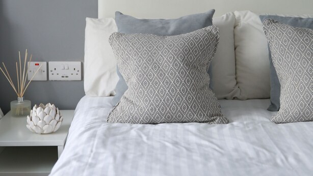 A neatly made bed with patterned pillows, a bedside table with a vase and reed diffuser, and a wall-mounted light fixture. - Greenmount Bed & Breakfast (Belfast)