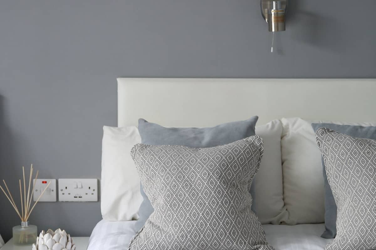 A neatly made bed with patterned pillows, a bedside table with a vase and reed diffuser, and a wall-mounted light fixture.