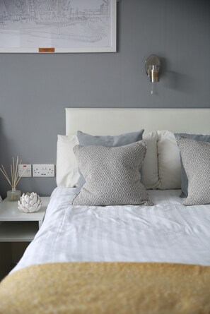 Un letto rifatto con cuscini decorati, un comodino con un vaso e un diffusore di fragranze, e un lampadario fissato al muro.