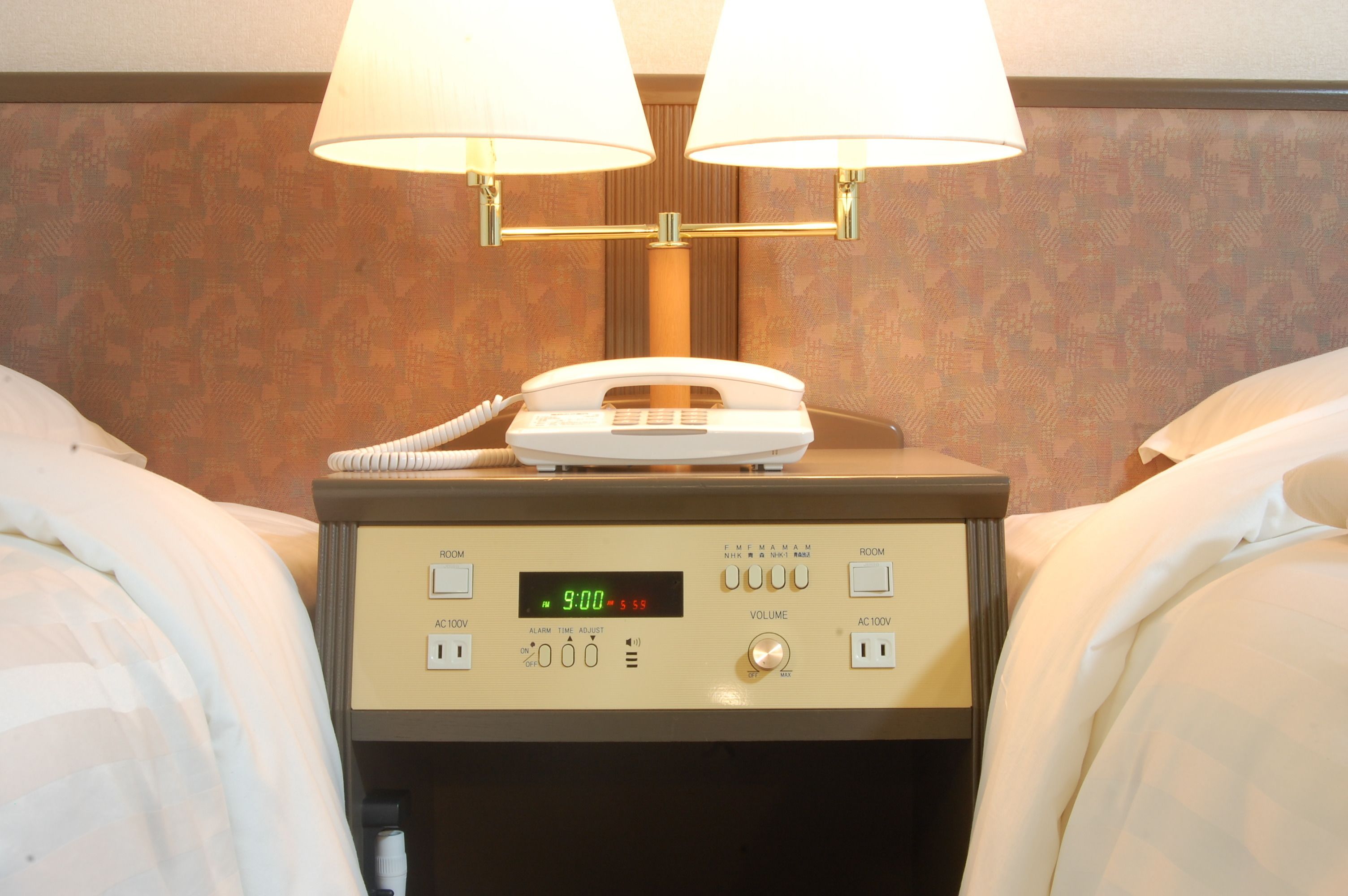 Une chambre d’hôtel dotée d’une table de chevet sur laquelle se trouvent un téléphone blanc, une horloge numérique et des boutons de commande pour régler les paramètres de la chambre.