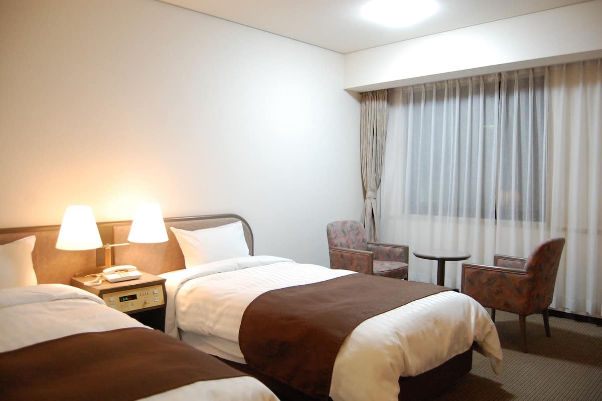 A hotel room with two beds, a nightstand with a phone, two lamps, a chair, a small table, and a large window with curtains.