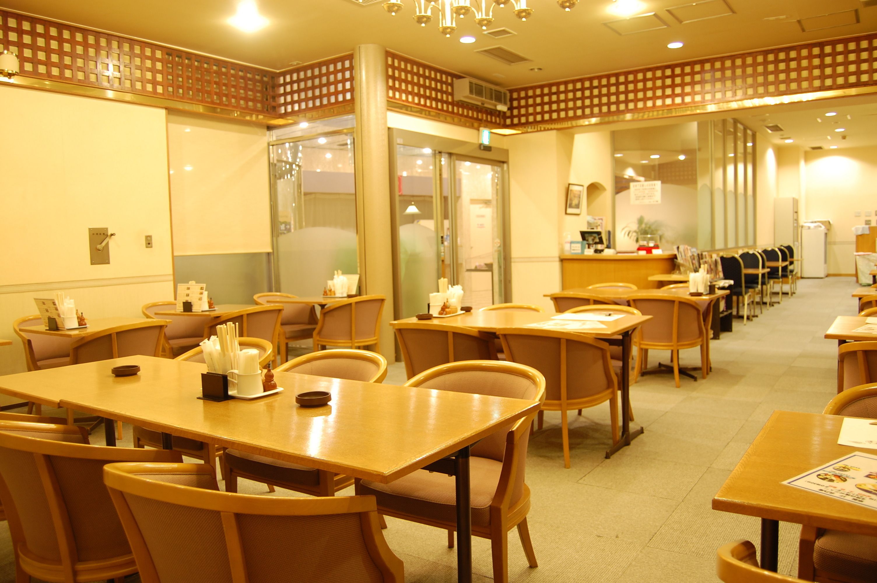 A hotel dining area with wooden tables and chairs, a reception desk, and a glass partition wall.