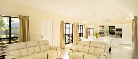 A modern living room with a beige sofa set, a wooden coffee table, and a kitchen area in the background.
