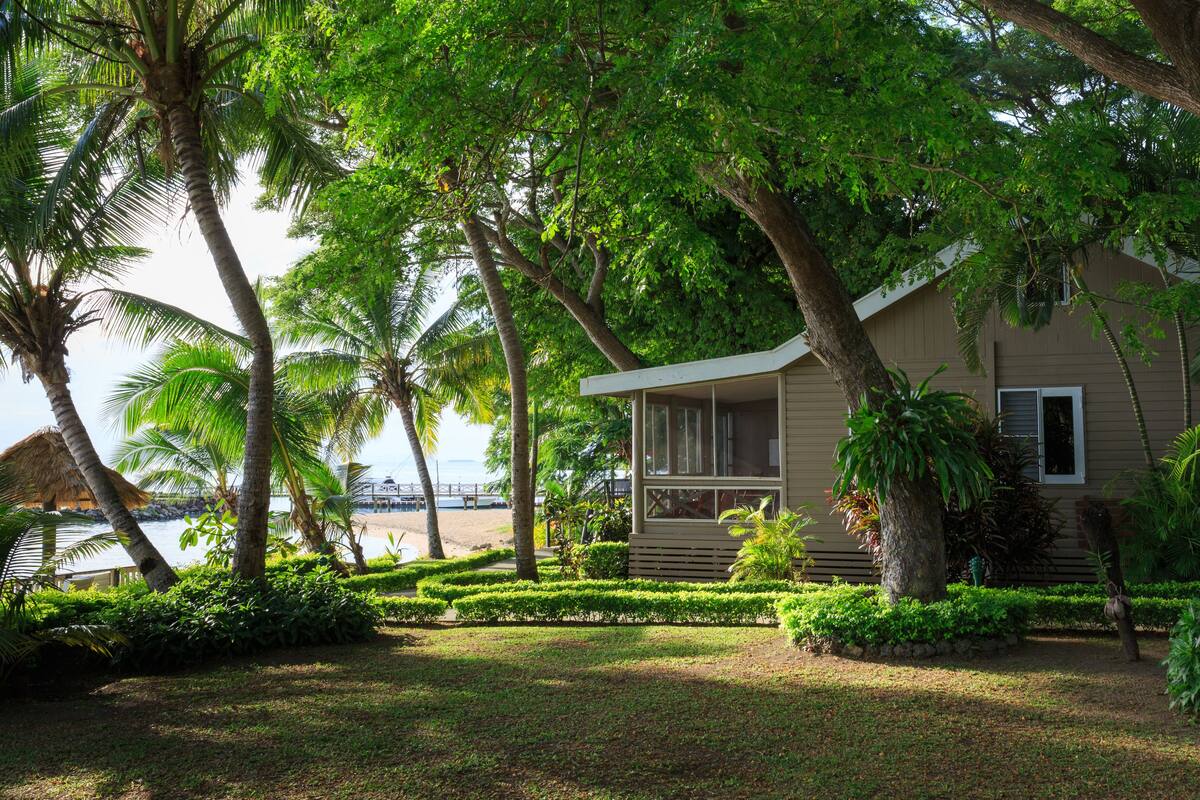 A bungalow with a porch, surrounded by lush greenery and palm trees, overlooking a beach.