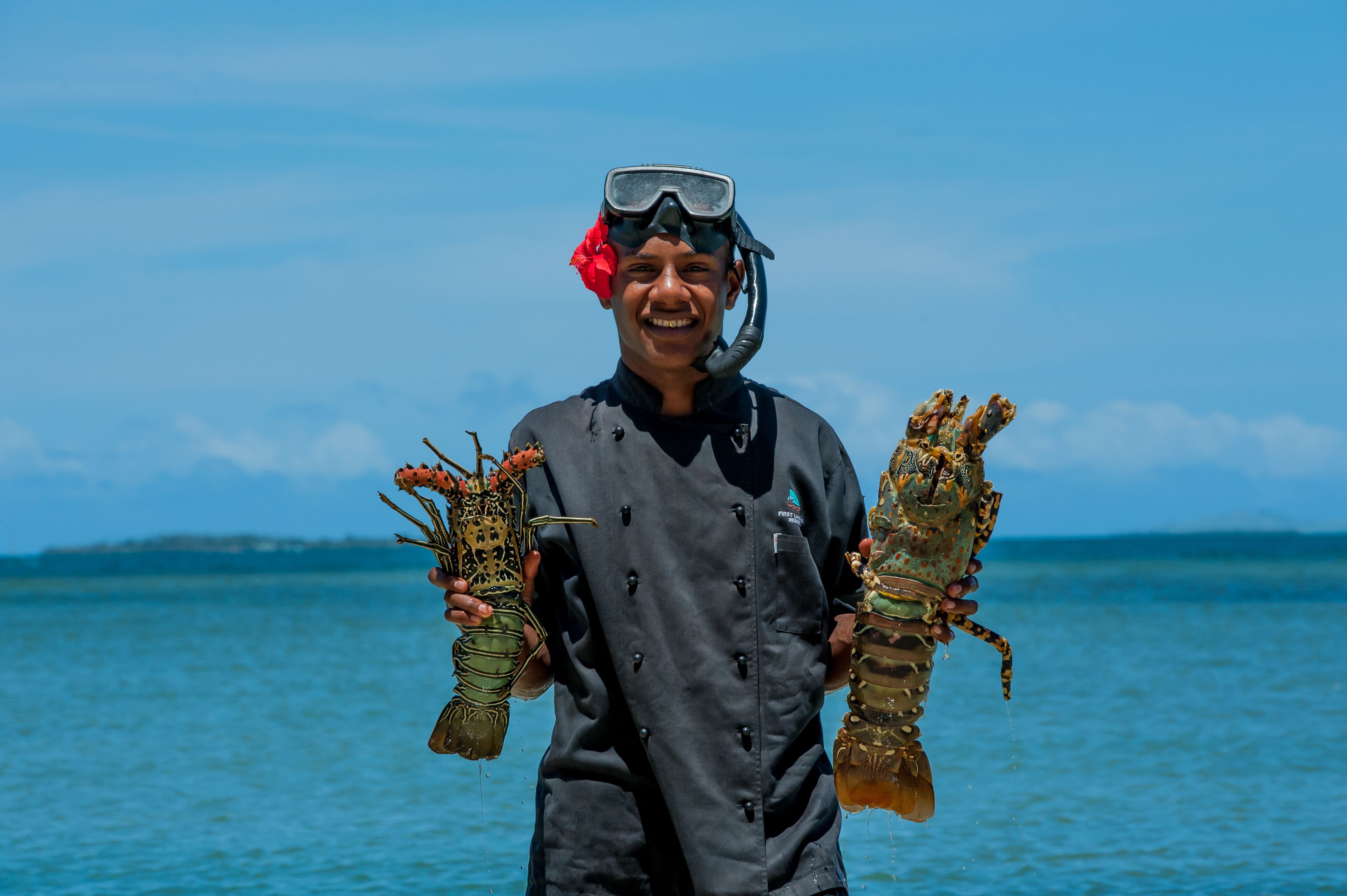 Una persona sosteniendo dos langostas, usando traje de buceo y snorkel, de pie junto a una masa de agua.