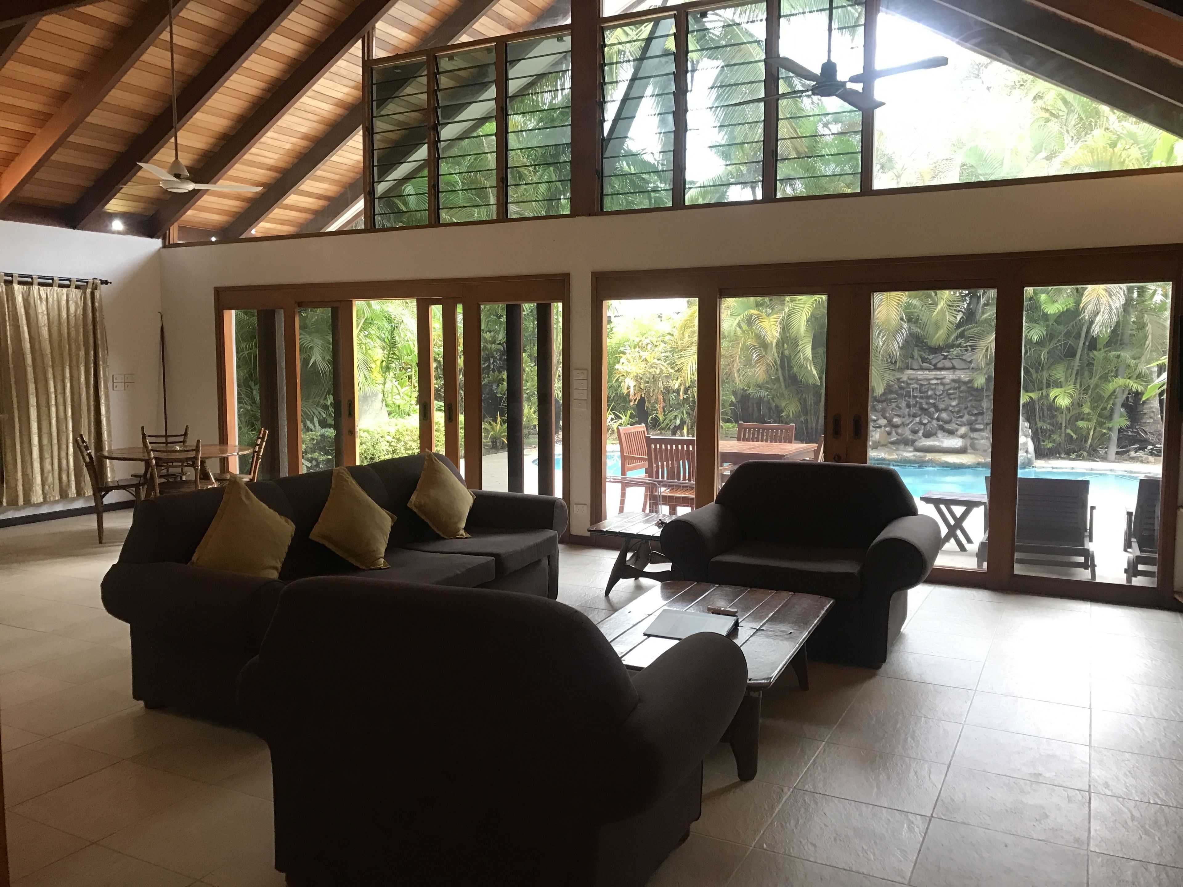 A spacious living area with a high ceiling, large windows, and a view of a pool and tropical garden.