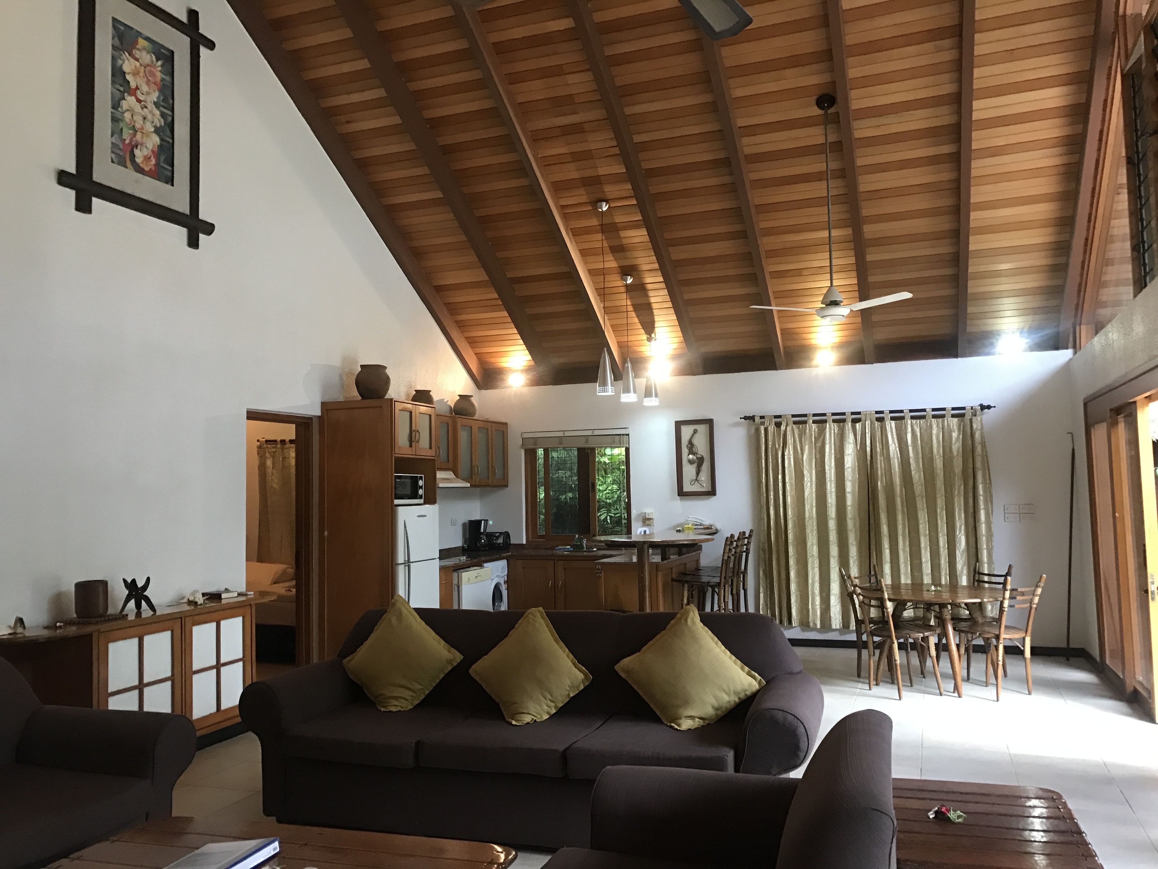 A spacious living area with a dark-colored sofa, wooden ceiling beams, a dining area with a table and chairs, and a kitchen with wooden cabinets.