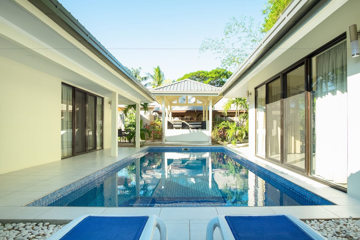 A swimming pool with lounge chairs, a covered seating area, and a view of a building with large windows and a tiled roof.