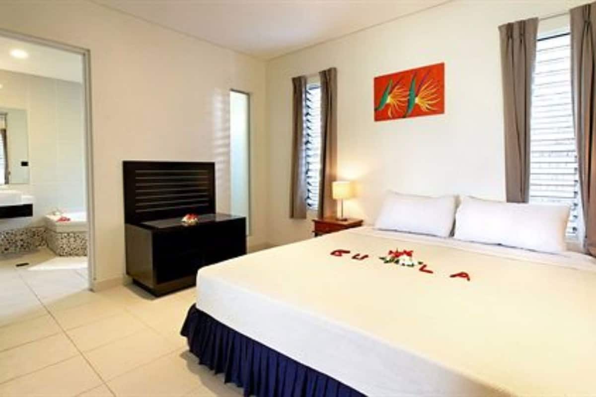 A hotel room with a bed, a bedside table, a nightstand, a mirror, a window with curtains, and a wall art piece.