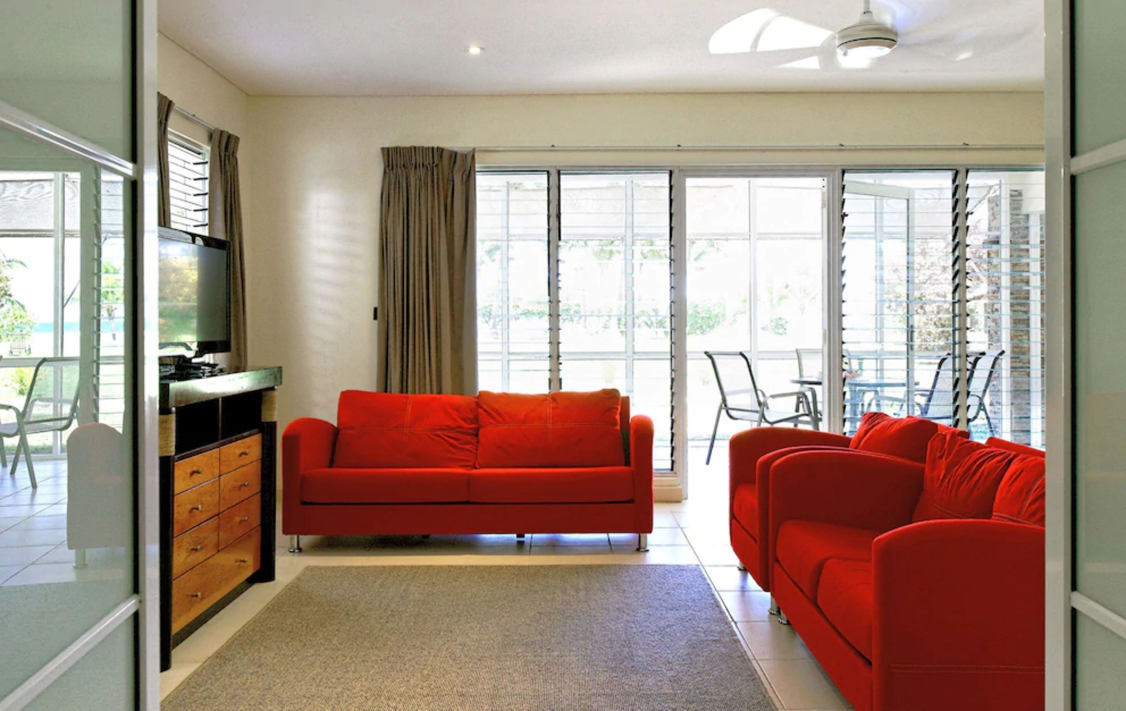 A modern living room with a red sofa, a TV on a wooden cabinet, and large windows with curtains.