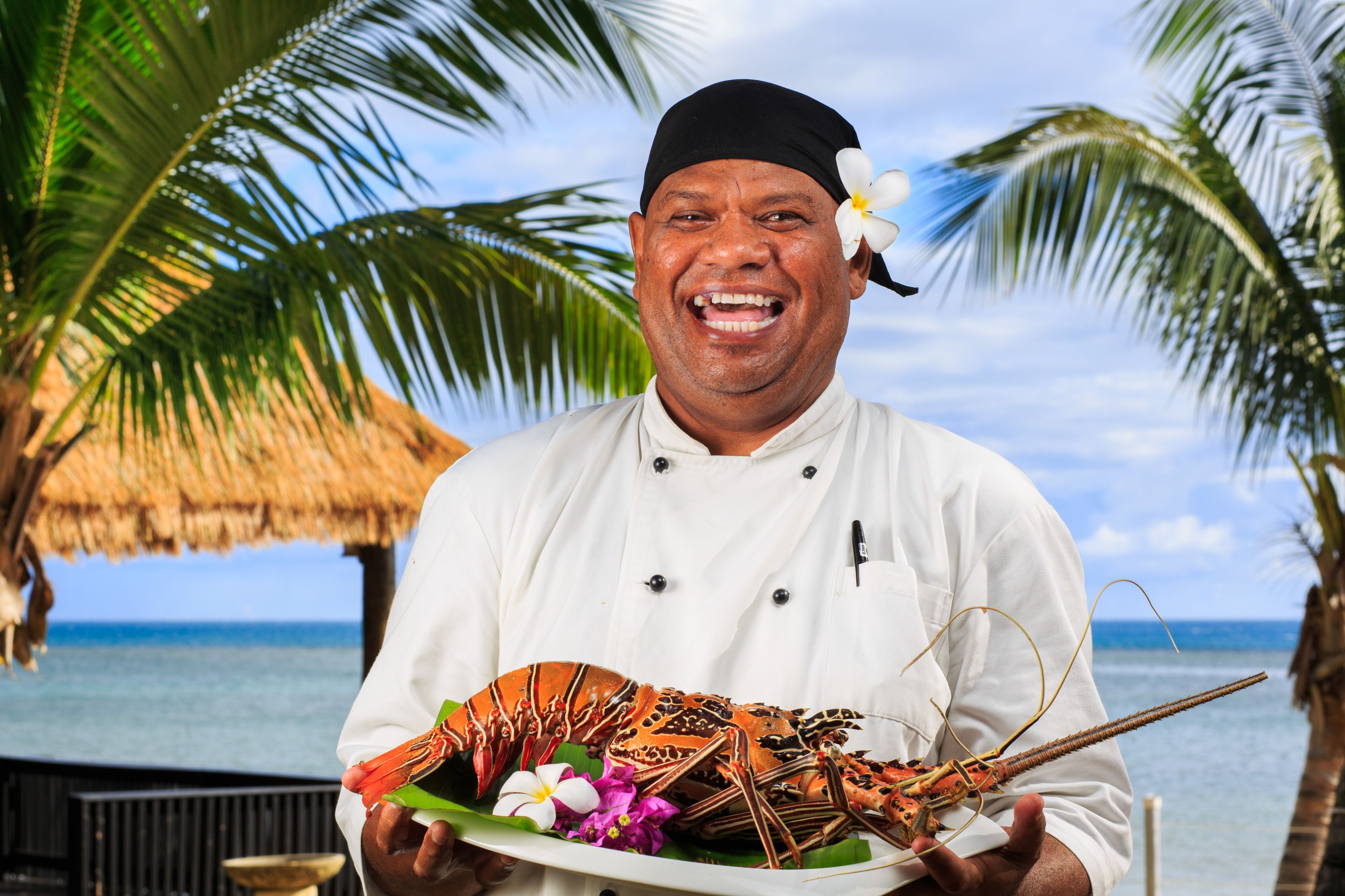 A chef holding a lobster on a plate with flowers, standing in front of a thatched structure and palm trees by the ocean.