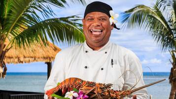 A chef holding a lobster on a plate with flowers, standing in front of a thatched structure and palm trees by the ocean.