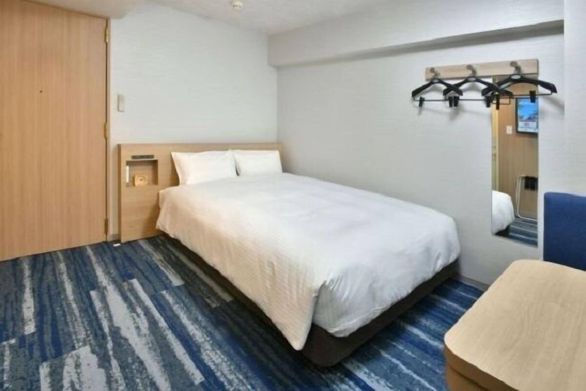 A hotel room with a large bed, a small bedside table, a chair, a coat rack, and a mirror.