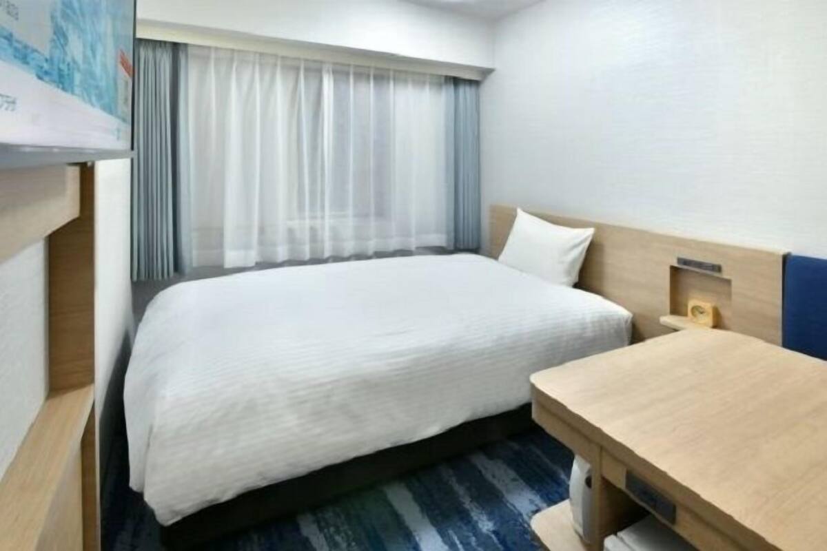 A hotel room with a single bed, a wooden nightstand, a small table, and a TV mounted on the wall.