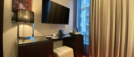 A hotel room with a flat-screen TV mounted on the wall, a desk with a telephone and a coffee pot, a small white ottoman, and a red carpeted floor.