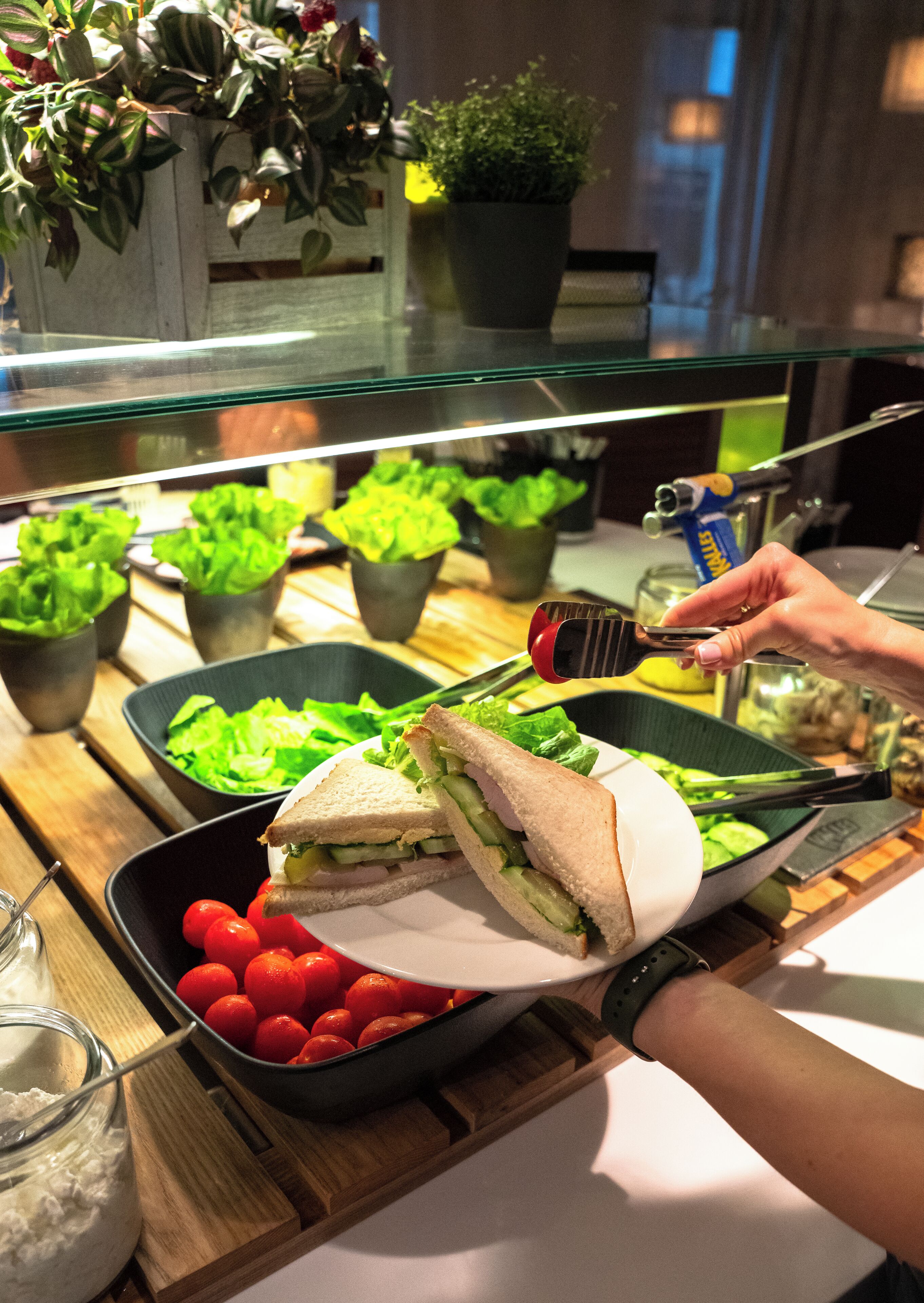 A sandwich being cut with tongs, surrounded by various vegetables and a bowl of cherry tomatoes.
