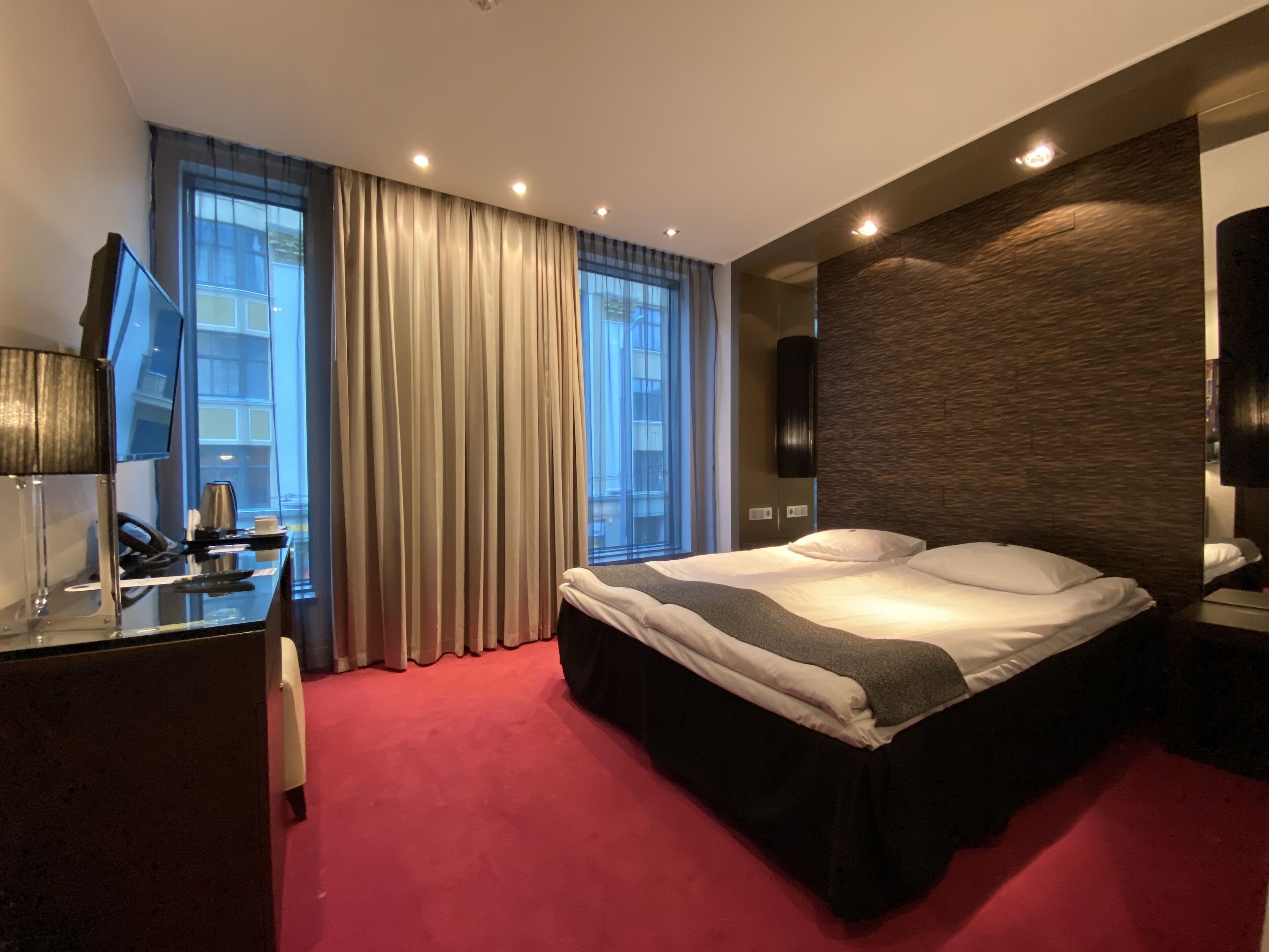 Una habitación de hotel moderna con una cama grande, un escritorio con computadora y lámpara, un televisor de pantalla plana y un ventanal con cortinas.