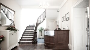 A hotel reception area with a wooden reception desk, a staircase with a black railing, and a chandelier. - Plas Hyfryd Hotel (Narberth)