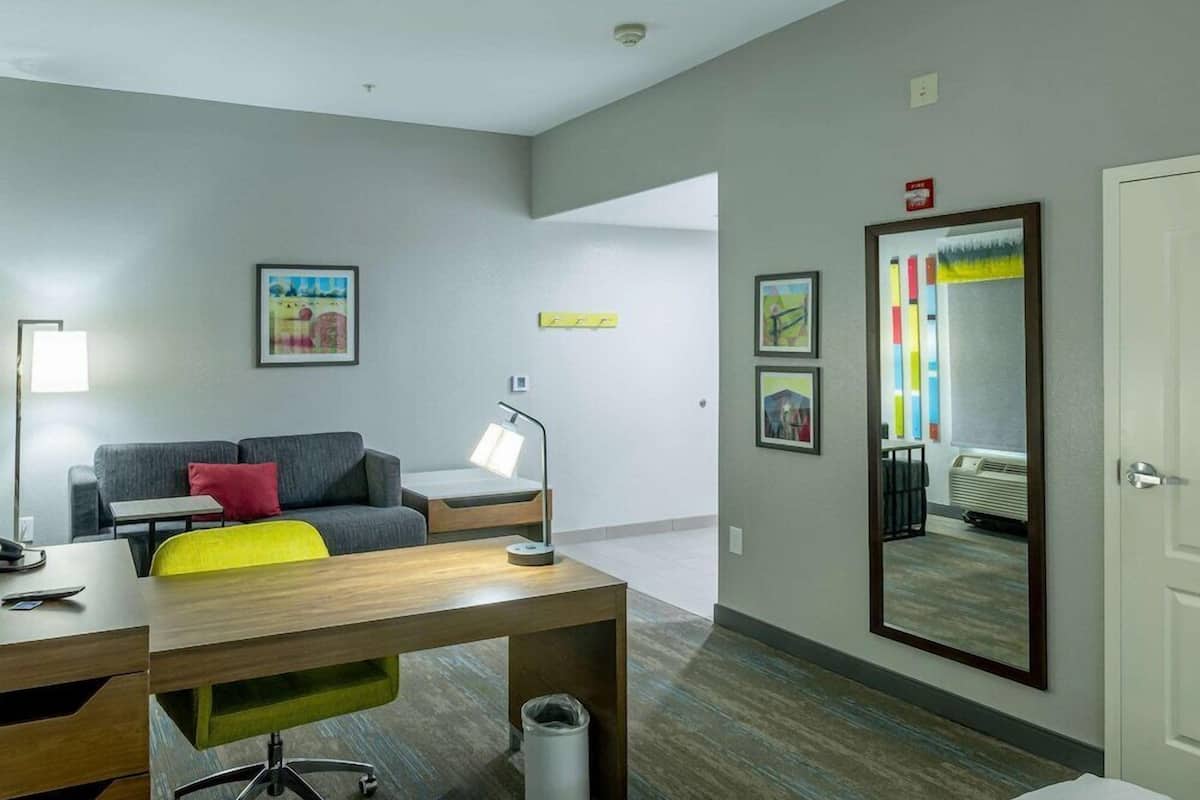 A modern hotel room with a desk, a sofa, a standing lamp, and a large mirror.