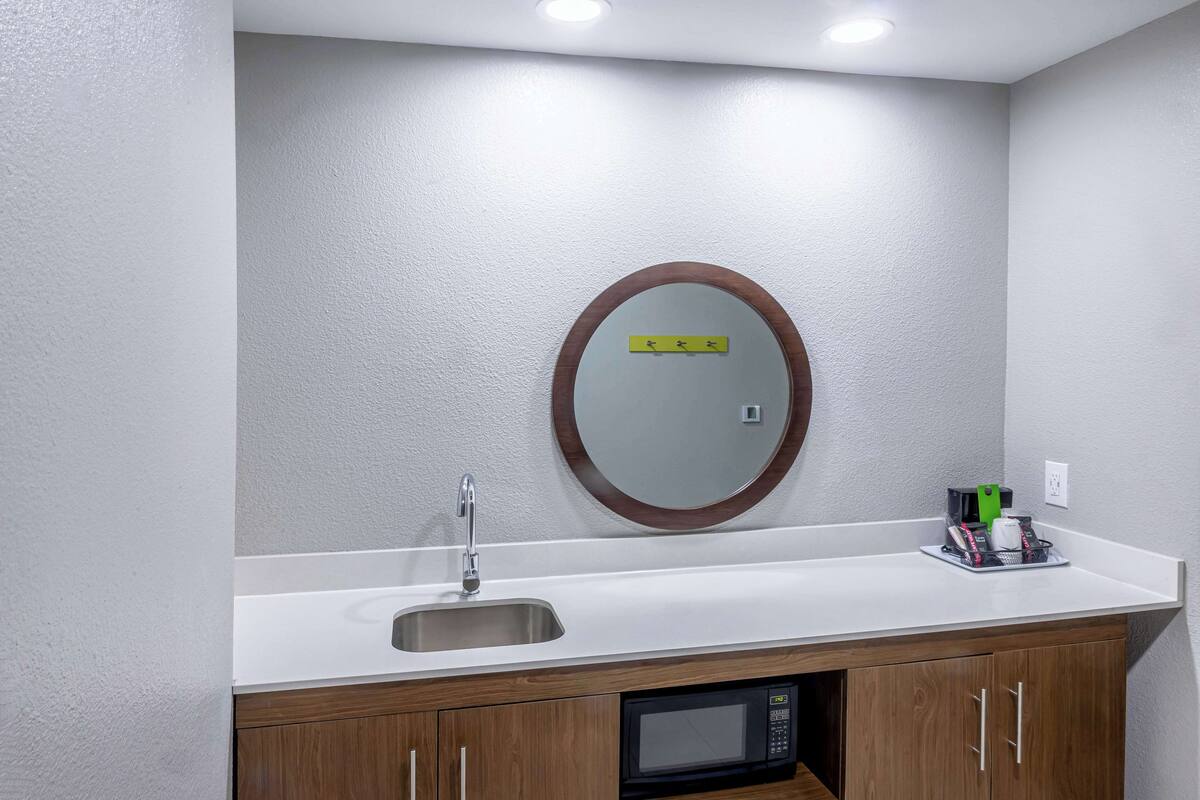 A modern bathroom with a white sink, a mirror with a wooden frame, a microwave, and a small shelf with toiletries.