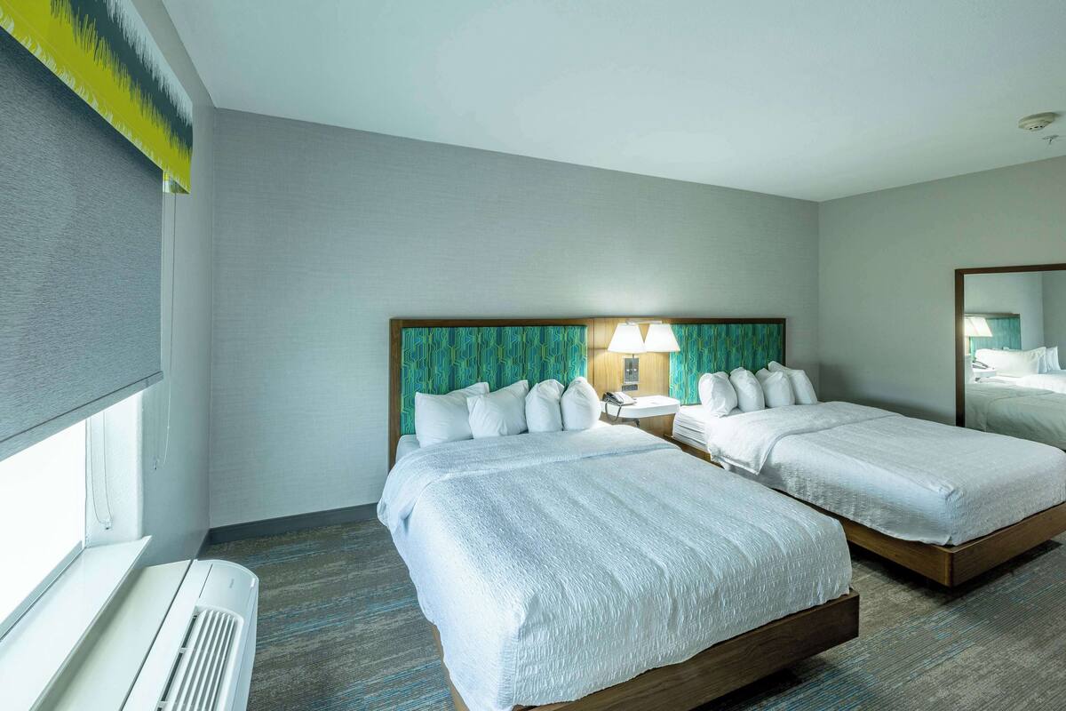 A hotel room with two beds, a window with blinds, a bedside table with a lamp, and a mirror on the wall.